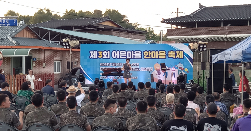 제3회 어은마을 한마음 축제 타이틀이 무대 벽면에 꾸며져 있고 군인들과 마을 사람들이 무대를 관람하고있다